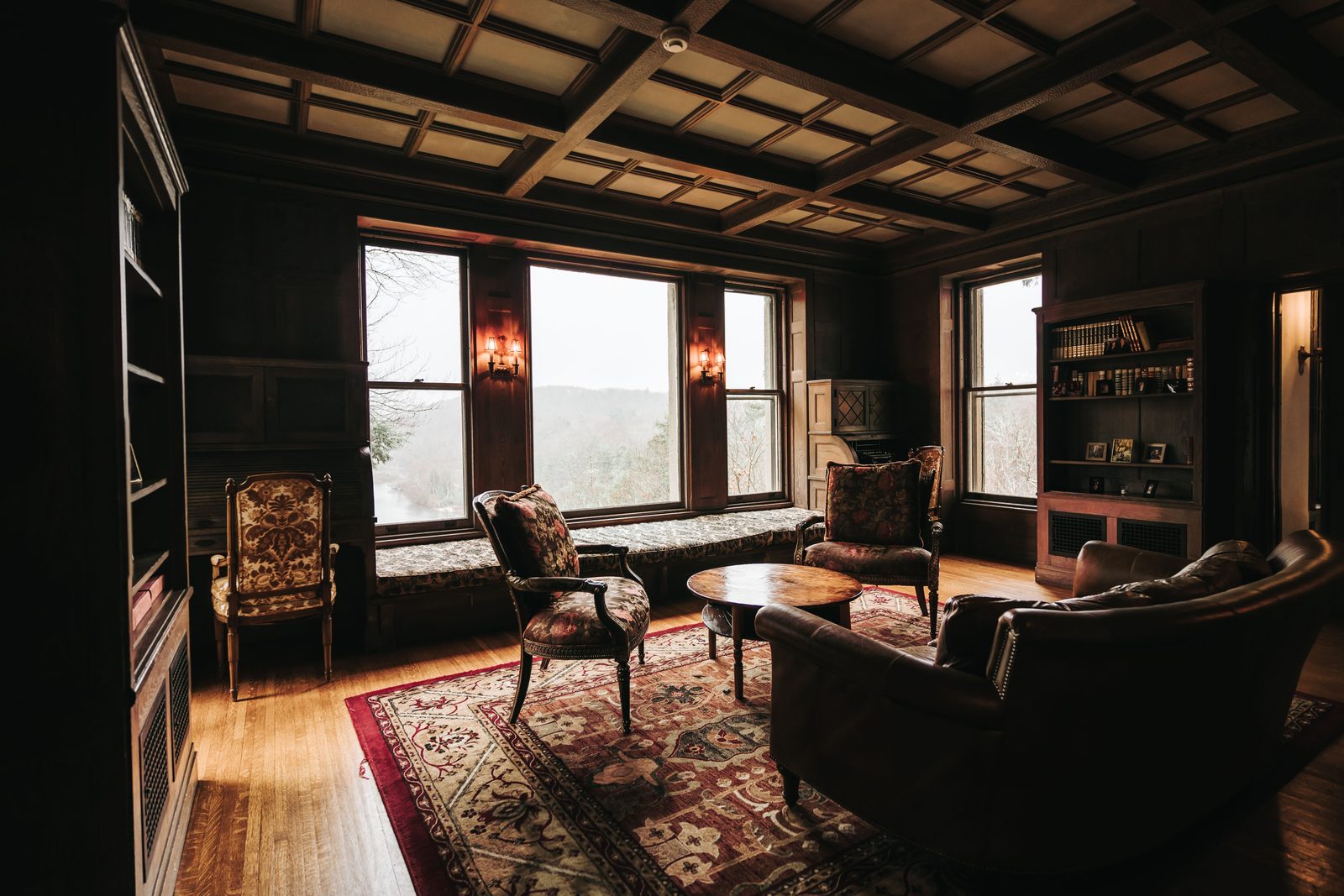 A dark wood-paneled library with a coffered ceiling, leather armchairs, a patterned rug and tall windows.
